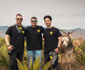 raicilla pkgd tours Three men in black shirts standing with a donkey in a field with mountains in the background