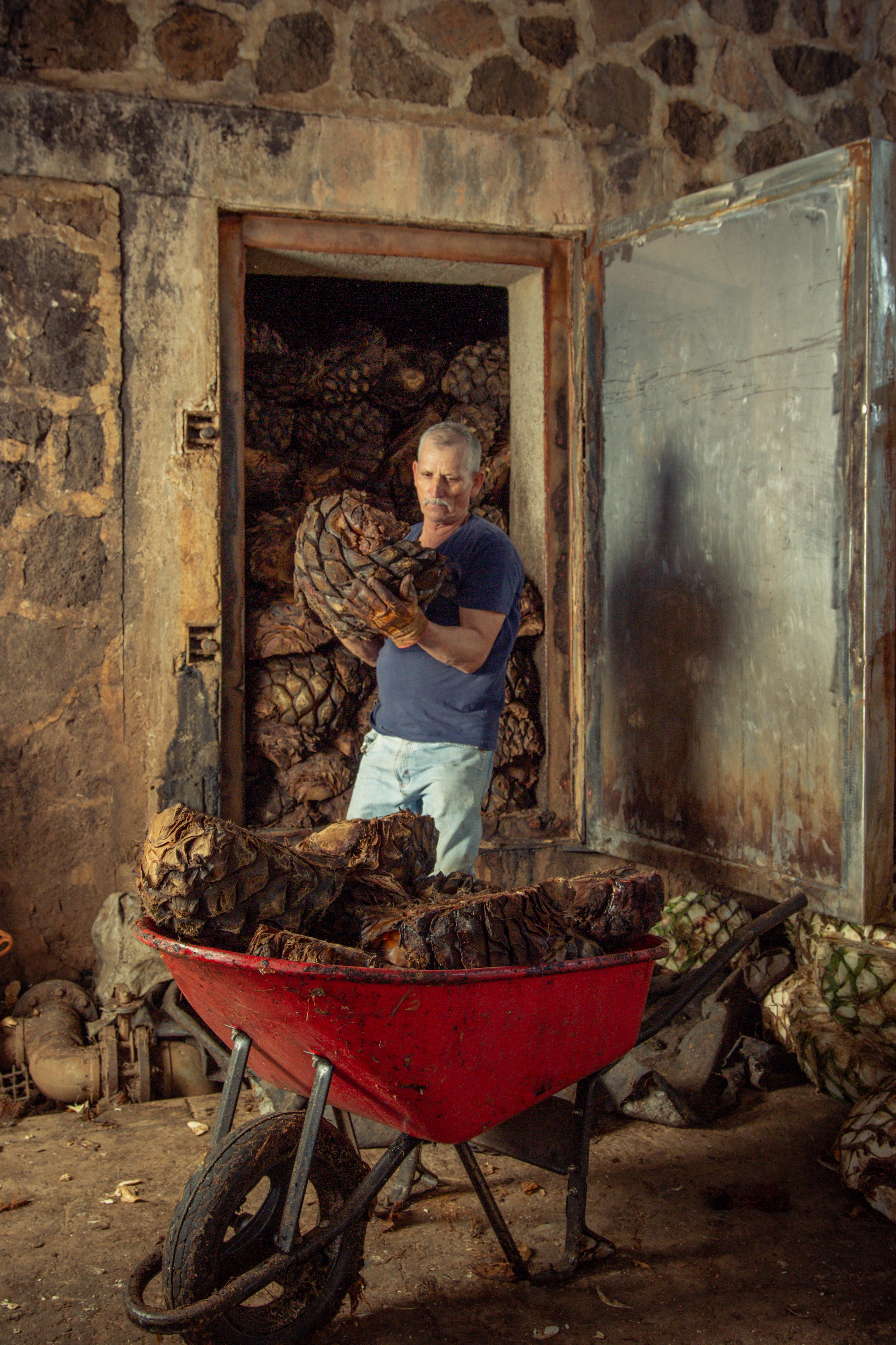 Man loading piñas into a red wheelbarrow in front of a stone building.
