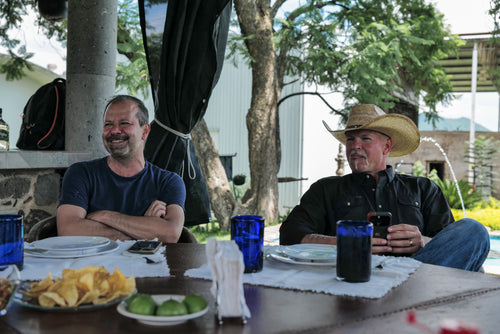 Two men sitting at a table outdoors with drinks and snacks.