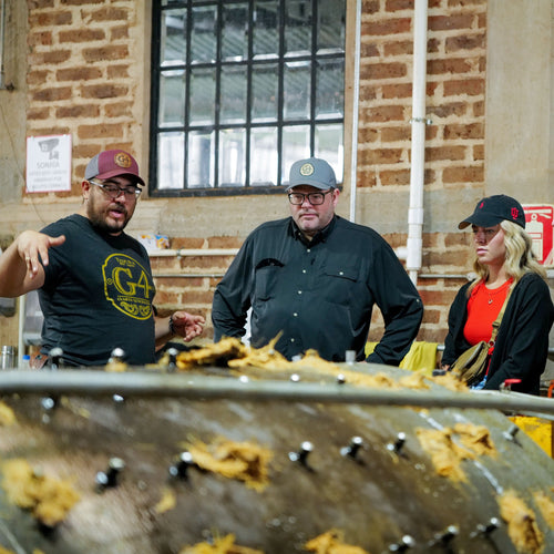 Three people standing near a large metal container with food items, against a brick wall.